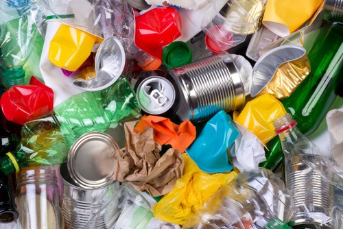 A close-up view of a mixture of discarded plastic and metal packaging materials, including empty aluminium cans, plastic bottles, and crumpled plastic wrappers, scattered across a surface. The aluminium cans are silver with visible pull-tabs, some partially crushed or dented, while the plastic bottles are in various colors such as green, clear, and yellow, with some caps attached. Crumpled paper and plastic wrappers in bright colors like red, yellow, orange, and blue are mixed among the larger items. The background shows more debris, suggesting a piled or cluttered scene typical of waste materials awaiting collection or removal. This image reflects common rubbish encountered in residential or commercial rubbish removal services, such as those provided by Garden Clearance Kings Cross in the local area around Kings Cross, London.