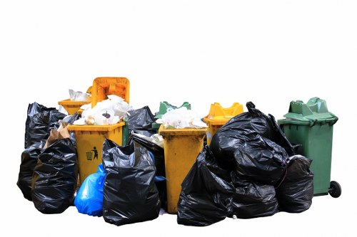 A collection of various rubbish bins and bags stacked together in an outdoor setting, including a yellow wheelie bin with a partially open lid, another yellow bin with a closed lid, a green bin on small wheels, and multiple black plastic rubbish bags of assorted sizes, some tied and others open with visible waste inside. The bins and bags are placed on a paved surface, with a plain white background that emphasizes the cleaning and waste disposal theme, suggesting a rubbish clearance task in a residential or commercial context. The scene reflects typical waste collection and disposal elements that Garden Clearance Kings Cross might handle in the Kings Cross area, highlighting the different container types and waste packaging used during rubbish removal services.