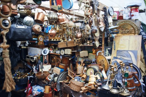 A densely packed outdoor display of assorted household and decorative items including vintage and antique metalware, brass and copper pots, vases, and teapots, along with clocks, picture frames, and small furniture pieces. The items are stacked and hung on a wooden structure, with some objects resting on a surrounding table or surface. The surface appears weathered, with a mix of shiny and tarnished finishes, primarily in metallic tones such as gold, silver, and bronze. Various textiles like fabrics and bags are also visible among the clutter, adding texture and colour diversity. There are no visible signs of modern machinery or disposal equipment, but the scene suggests a secondhand or antique market stall that could be part of a garden clearance in Kings Cross or nearby London suburbs. The background hints at an outdoor setting, likely a market or fair, with natural light illuminating the objects and casting subtle shadows, contributing to a busy, cluttered atmosphere typical of secondhand sales or clearance operations undertaken by Garden Clearance Kings Cross.