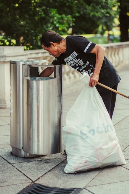 A sanitation worker wearing a blue uniform and an orange high-visibility vest is standing on a cobblestone street beside a white rubbish collection truck, which has its rear compactor open and partial