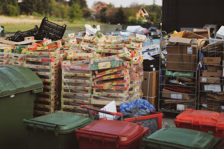 A large pile of mixed household and commercial waste is overflowing from several open and closed rubbish bins located on a paved area in front of a retail or commercial building. The waste includes fl