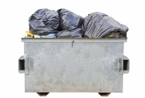 A large grey industrial skip bin filled with black rubbish bags and miscellaneous waste materials, situated outdoors on a paved area likely near a residential or commercial property in the Kings Cross area. The bin's surface shows slight weathering and minor stains, with two handles visible on each side for lifting. The rubbish bags are piled on top of each other, some slightly bulging, indicating they are full. The background includes a minimal view of a paved driveway or street, with natural daylight illuminating the scene, emphasizing the utilitarian purpose of waste collection and clearance services provided by Garden Clearance Kings Cross. The image reflects a typical scene of rubbish awaiting removal as part of a garden clearance or waste disposal service in the local area, featuring straightforward, practical waste storage with no additional objects present in the frame.