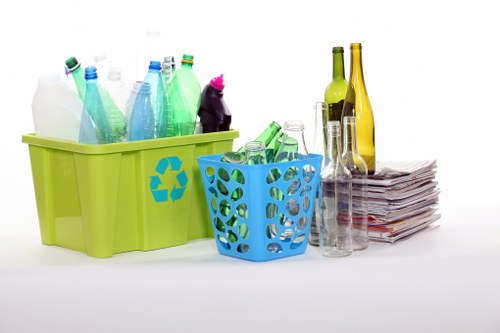 A collection of empty glass bottles and plastic containers in two waste bins – a larger green bin with a recycling symbol and a smaller blue bin – set against a plain white background. The green bin contains various plastic bottles of different shapes and colors, including blue and clear, with some bottles standing upright and others leaning. To its right is a blue basket filled with additional green plastic bottles, some lying on their sides. Adjacent to the bins is a neatly stacked pile of newspapers and magazines, with some clear glass bottles and tumblers placed nearby. The scene emphasizes waste segregation and recycling practices suitable for residential or commercial rubbish removal services in the Kings Cross area, with the objects arranged on a flat surface that highlights environmental responsibility in waste disposal and recycling efforts.